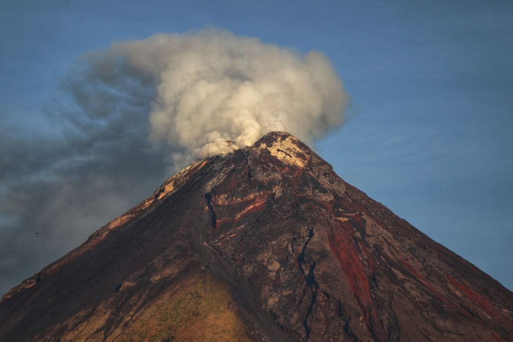 Tall bare conical volcano erupting with white heavy ash clouds under clear blue sky