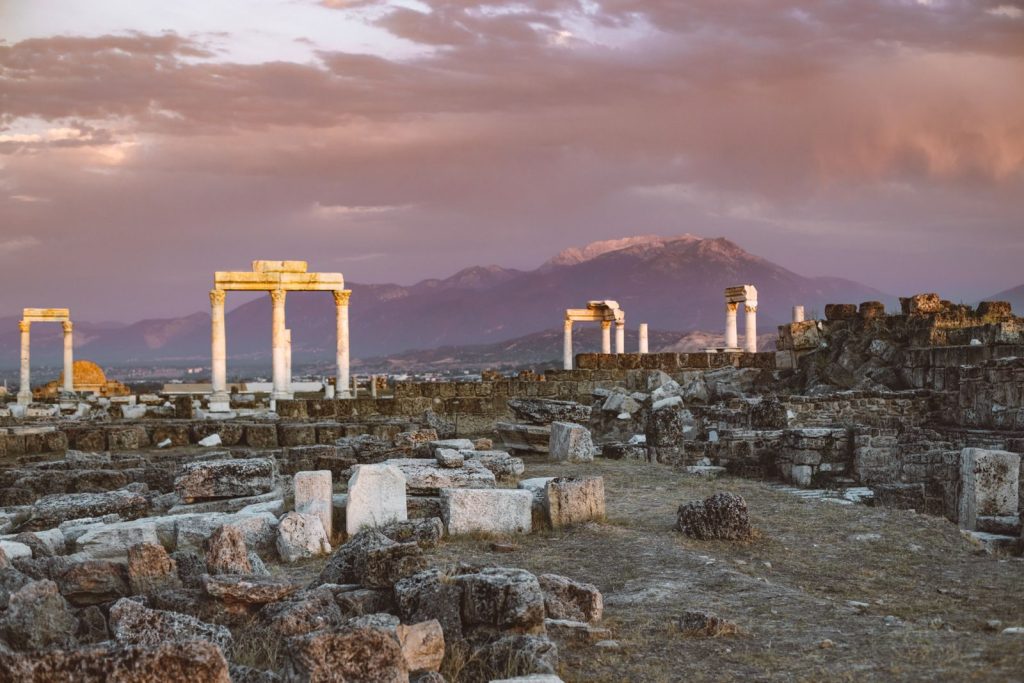 Explore ancient stone ruins and columns against a mountain backdrop during a stunning Turkish sunset.