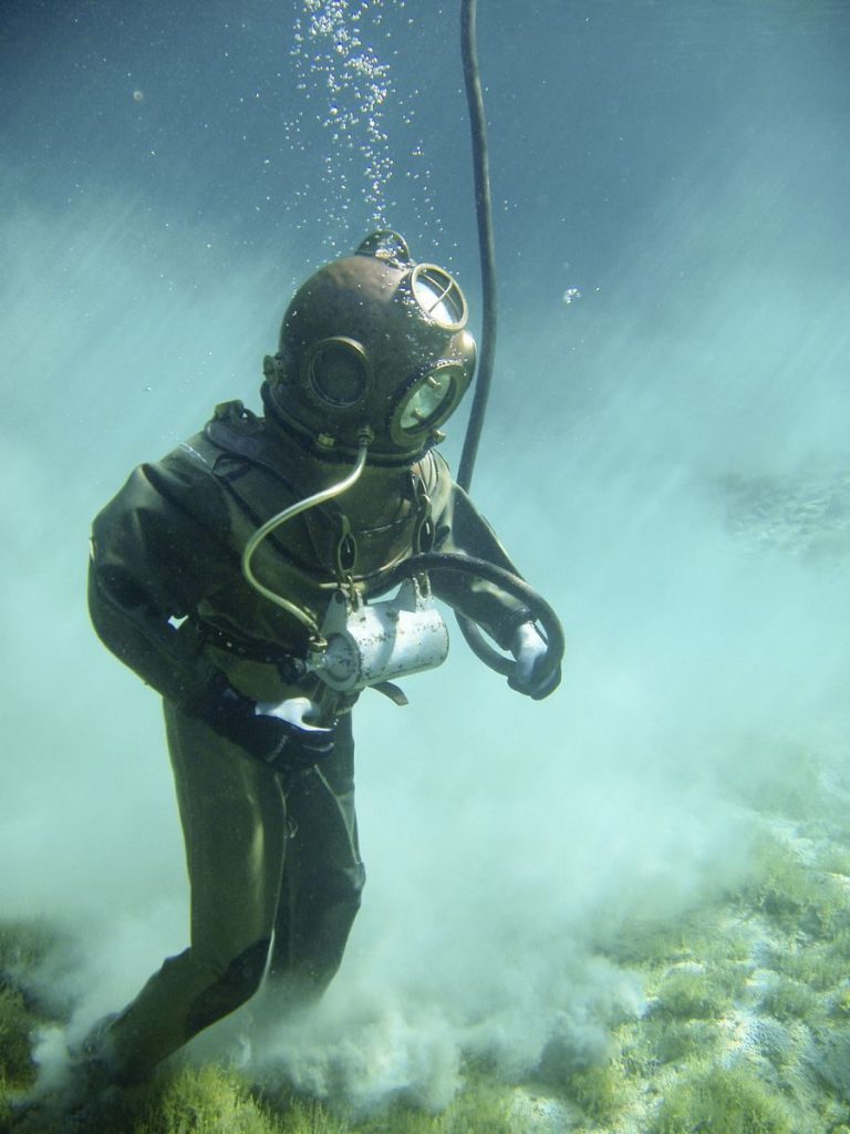A vintage diver in a classic suit explores underwater, surrounded by clouds of sand.