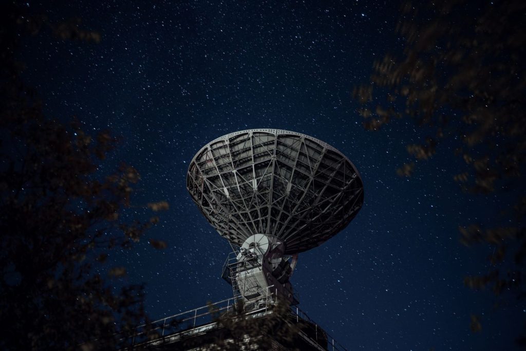 From below of blue starry sky over radio telescope and trees with leaves