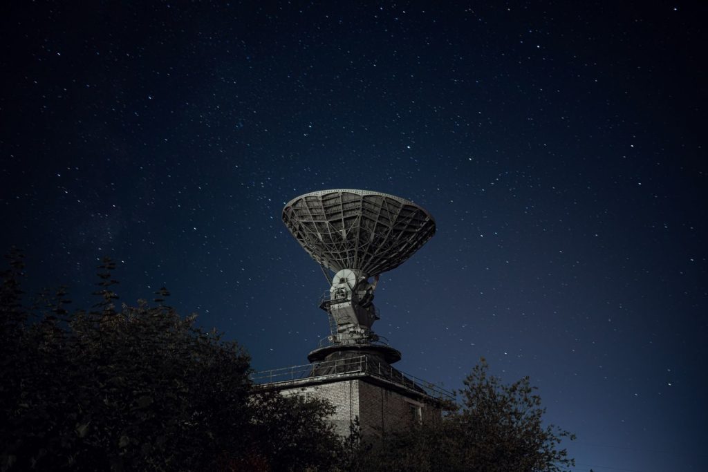Radio telescope and green lush trees under blue sky with stars at night