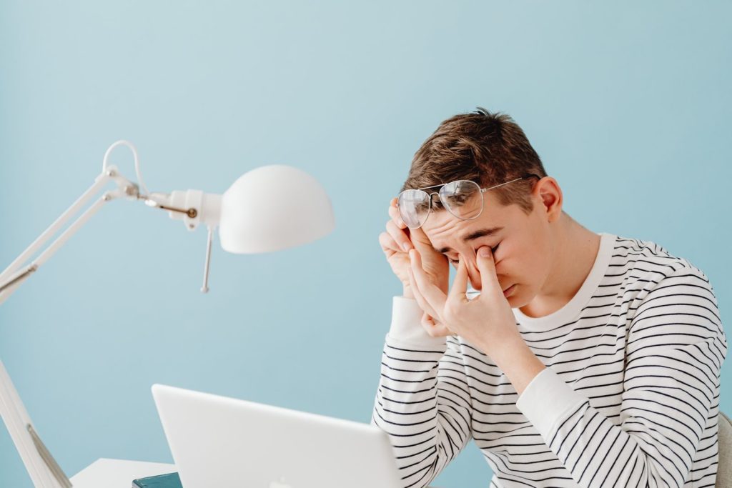 A young man rubs his eyes while working on a laptop, looking tired under a bright desk lamp.