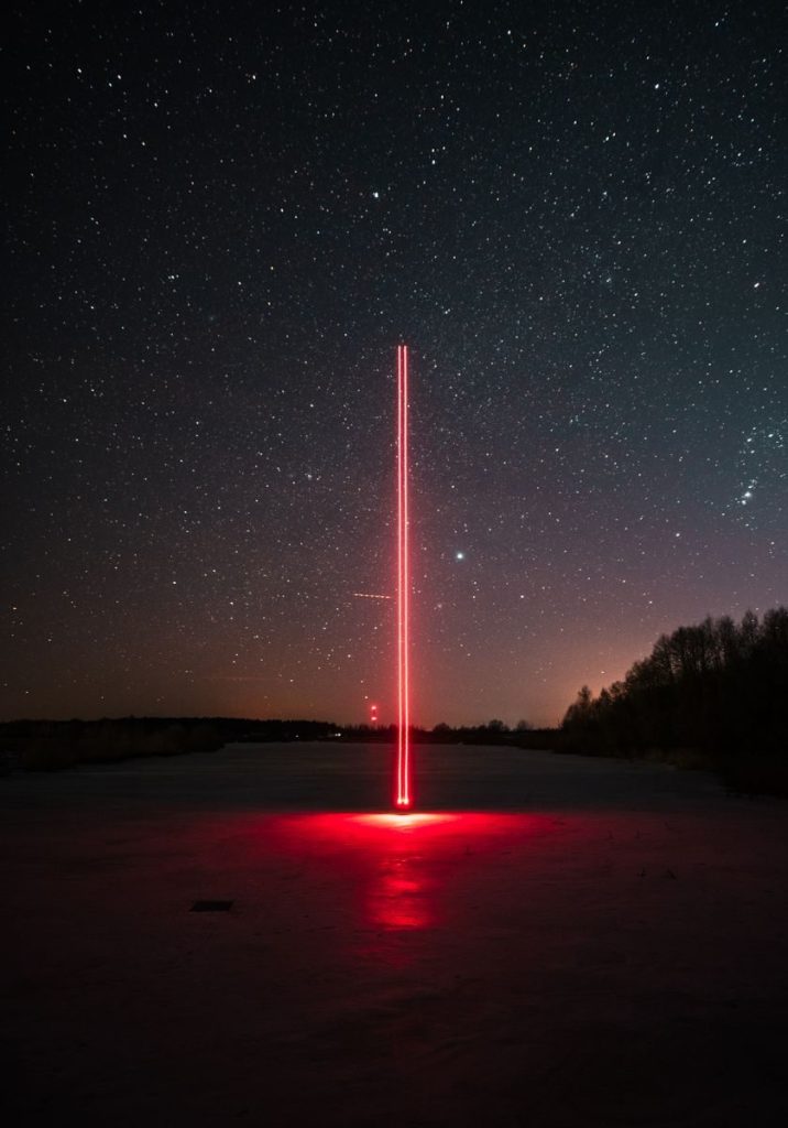 A striking red laser beam against a clear, starry night sky in a rural setting.
