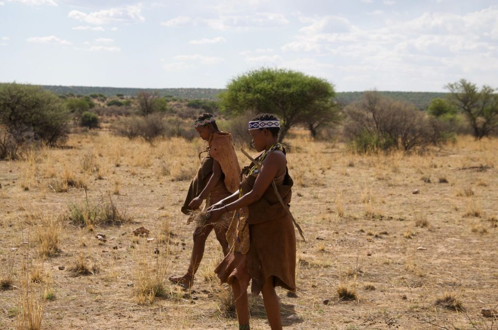 Two tribal members in traditional attire walking across a dry landscape surrounded by sparse vegetation.