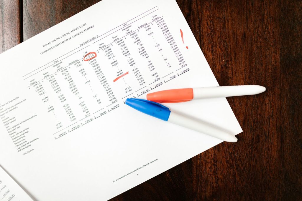 Top view of financial document with red marks and pens on wooden table surface.