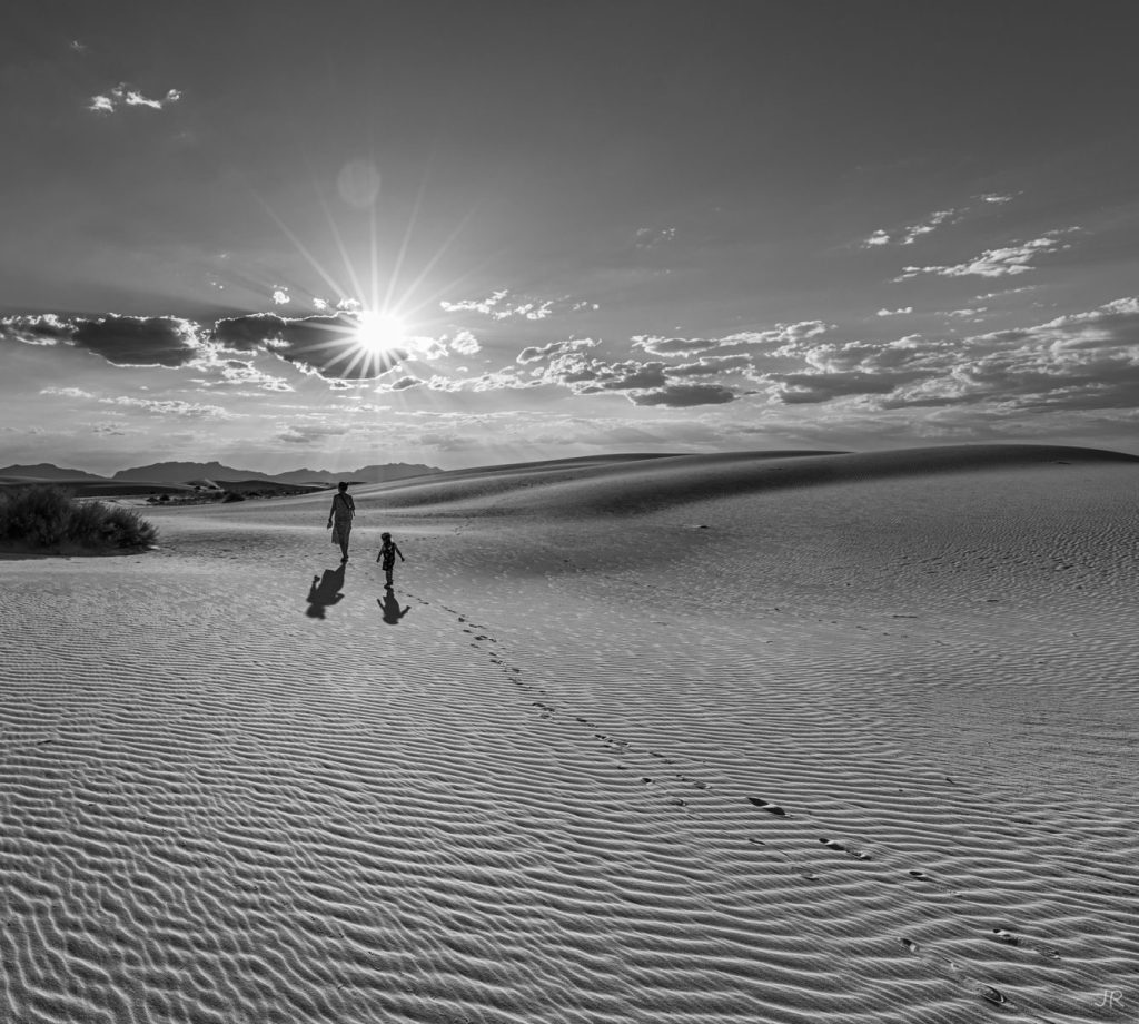 Silhouettes walking across White Sands desert at sunset, with dramatic shadows and sun flares.
