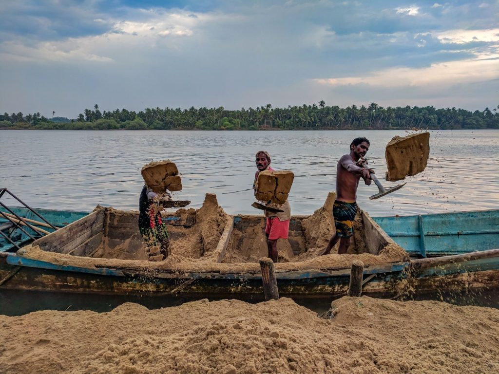 Three men dredging sand from a canoe on a peaceful lake with lush greenery in the background.