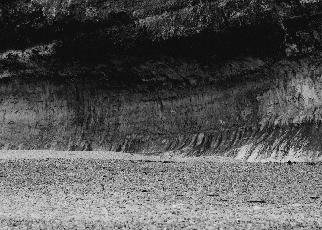 A cliffside overlooks a gravelly ground in monochrome.