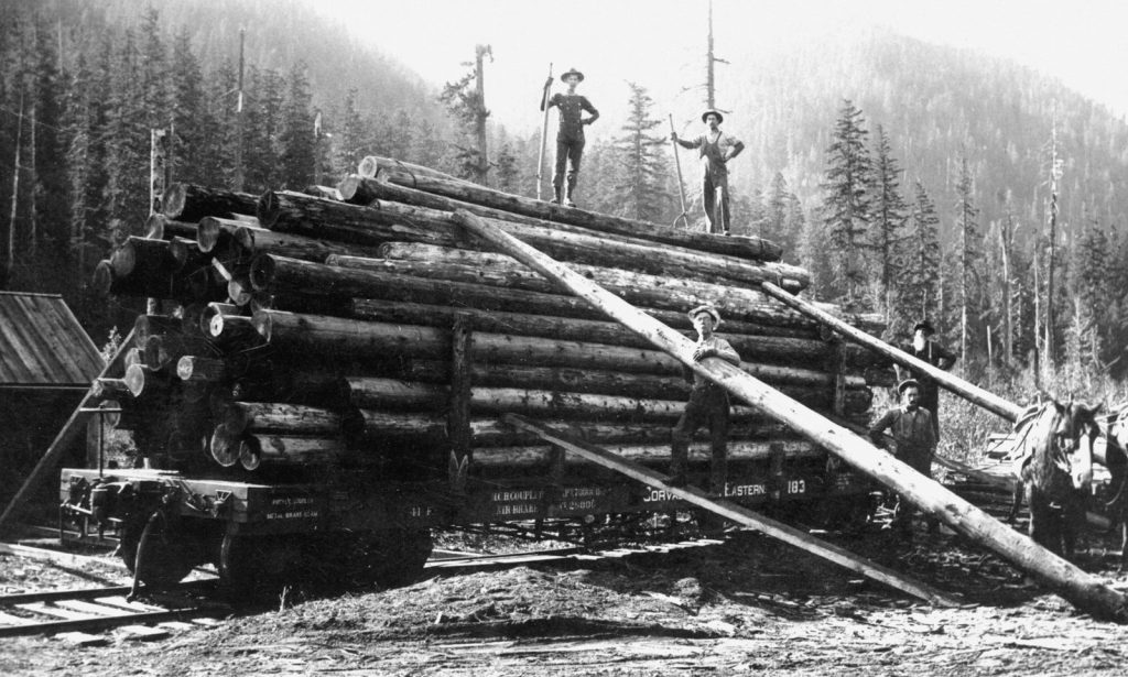Two men stand atop a loaded log cart with horses.