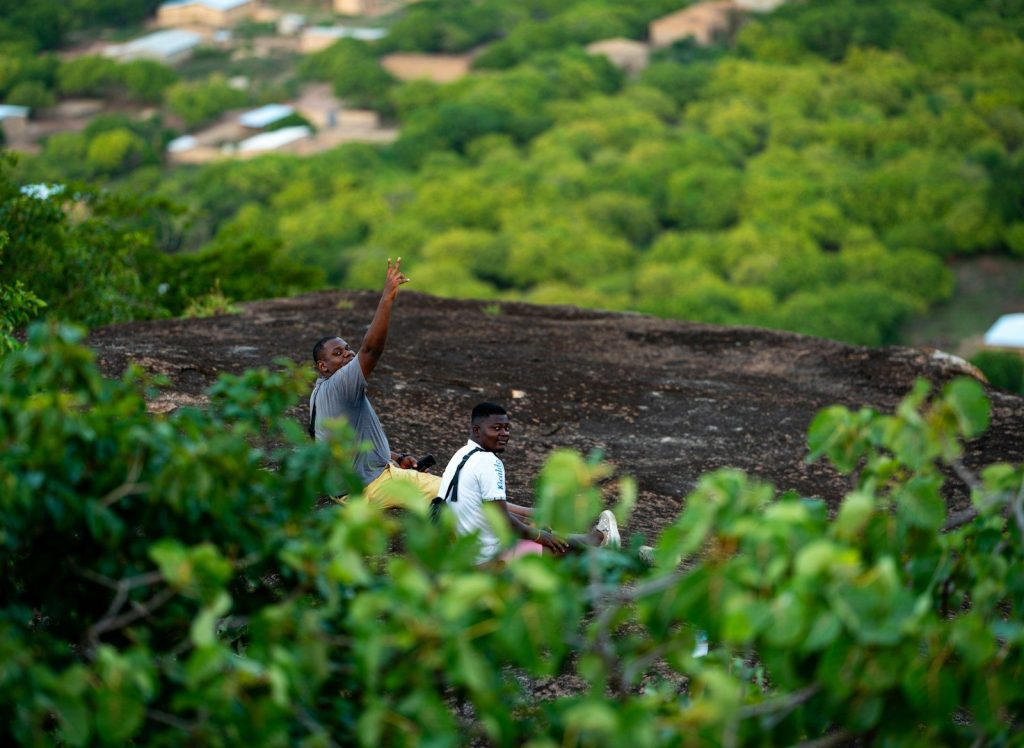 Man raising arm on rocky hill surrounded by green foliage.
