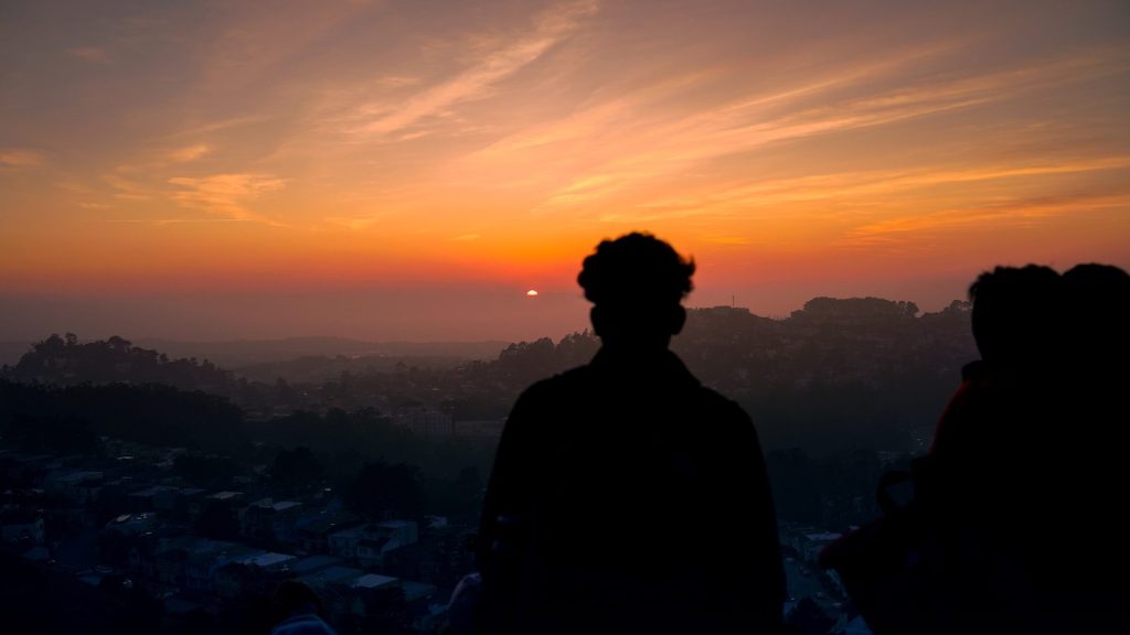 Silhouetted figures watch a vibrant sunset over hills.