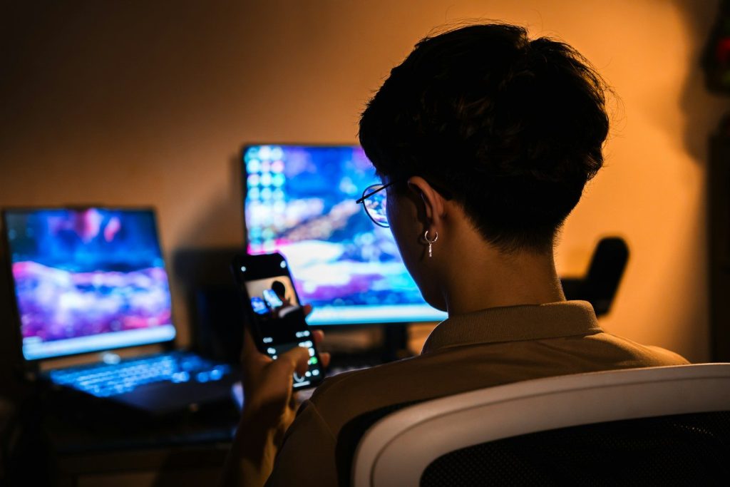 Person uses smartphone with glowing computer screens in dark room.