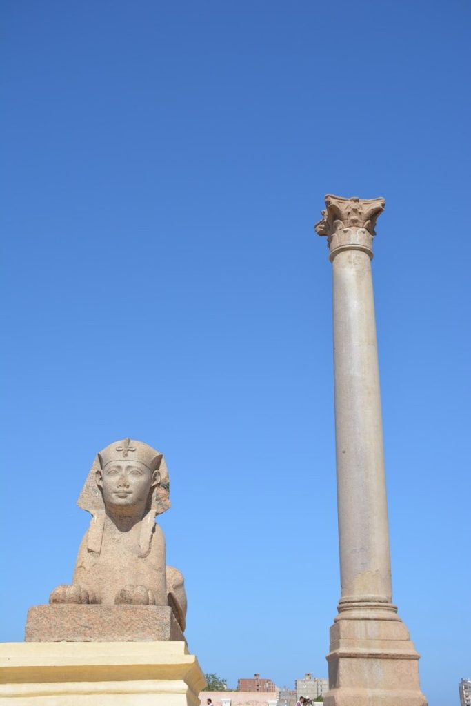 Ancient sphinx statue and roman column against a blue sky.