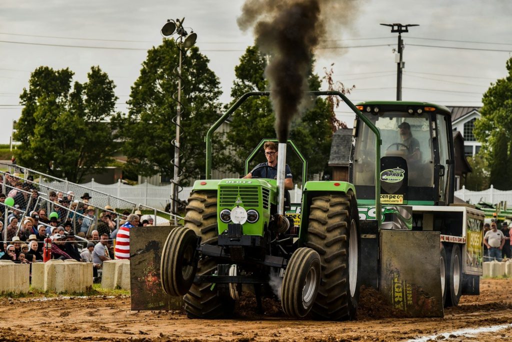 Powerful green tractor pulling sled, spewing black exhaust smoke.
