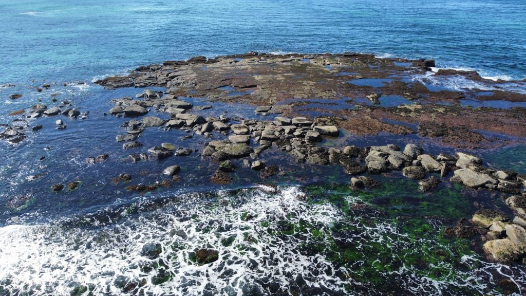 Aerial view of a rocky reef in clear blue ocean.