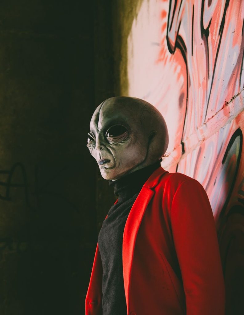 Alien figure wearing a red jacket stands against graffiti in an urban setting.
