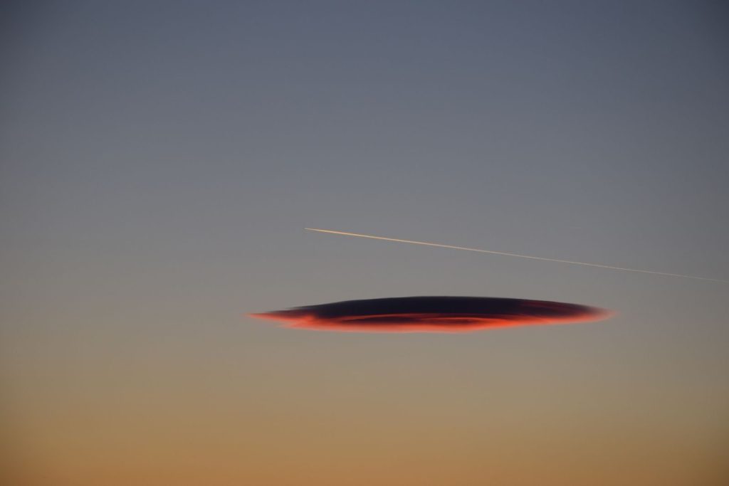 Lenticular cloud and chemtrail seen against a vibrant sunset sky, creating a dramatic scene.