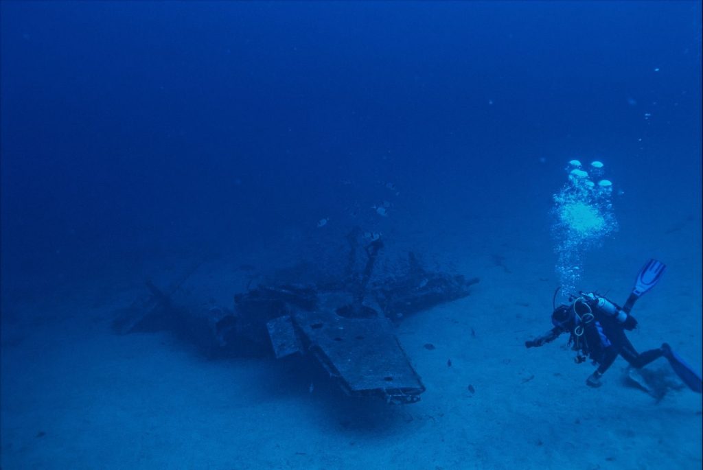 Unrecognizable person with oxygen tank emitting bubbles and swimming towards remains of sunken plane in blue sea water