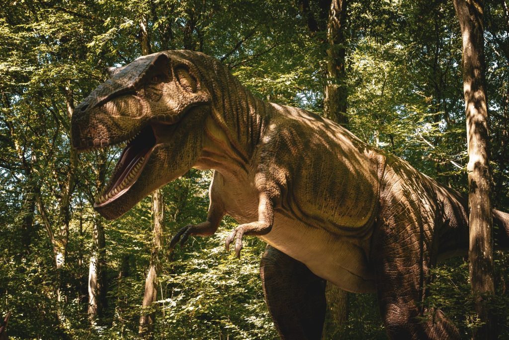 A life-sized dinosaur model stands in a sunlit, dense forest, creating a prehistoric atmosphere.