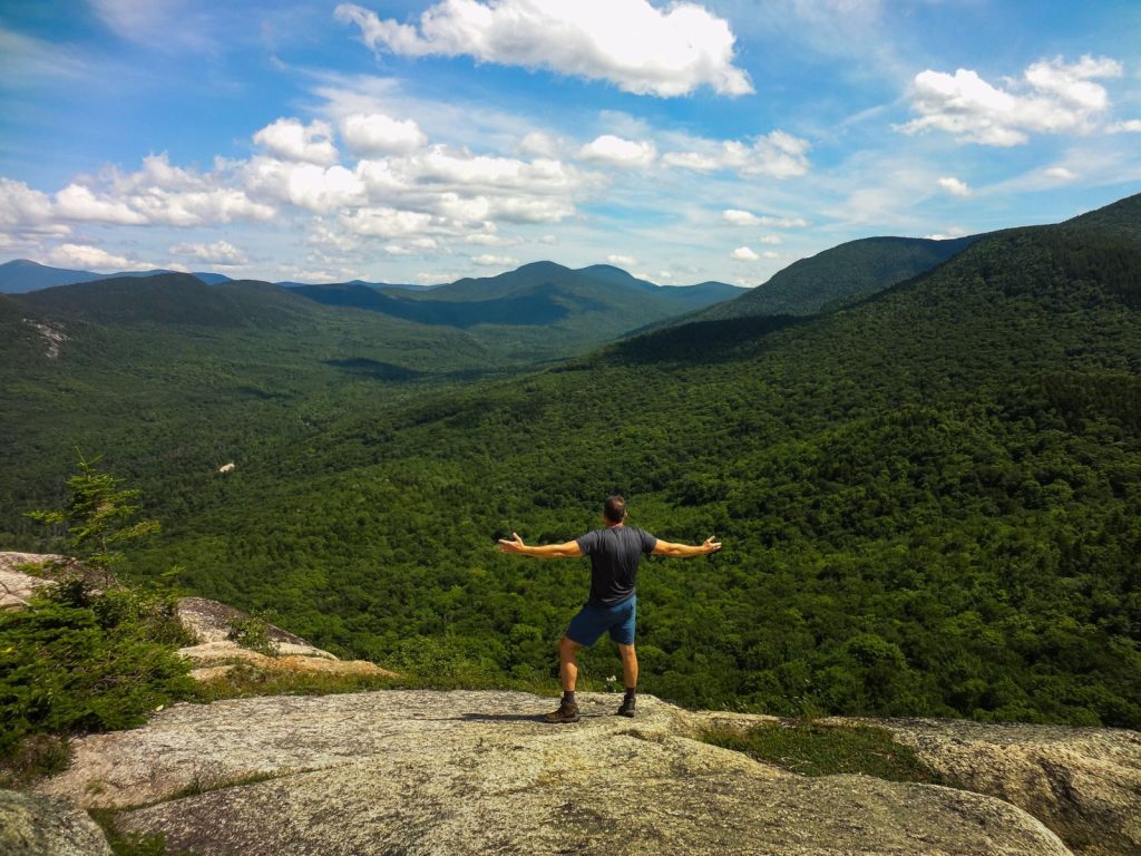 Hiker embracing nature on a rocky cliff, overlooking lush valleys in White Mountains, NH.