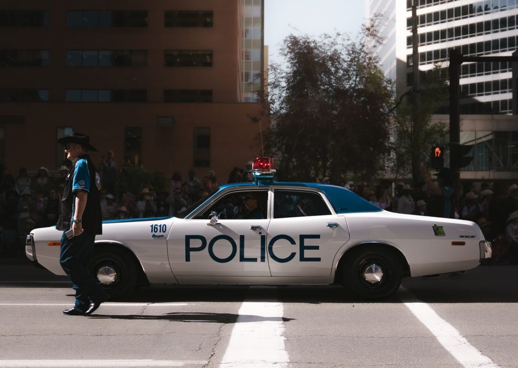 A vintage police car is featured in an urban parade with a crowd watching under a clear sky.