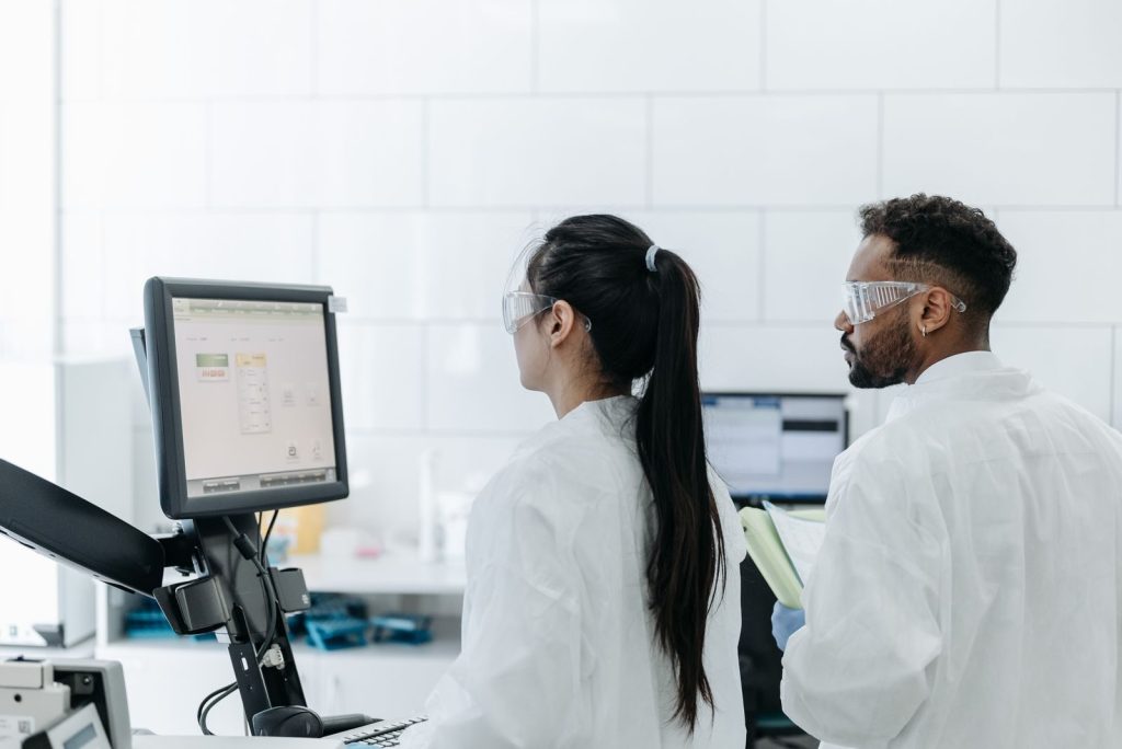 Two scientists wearing lab coats and goggles analyze data on a computer in a modern laboratory.